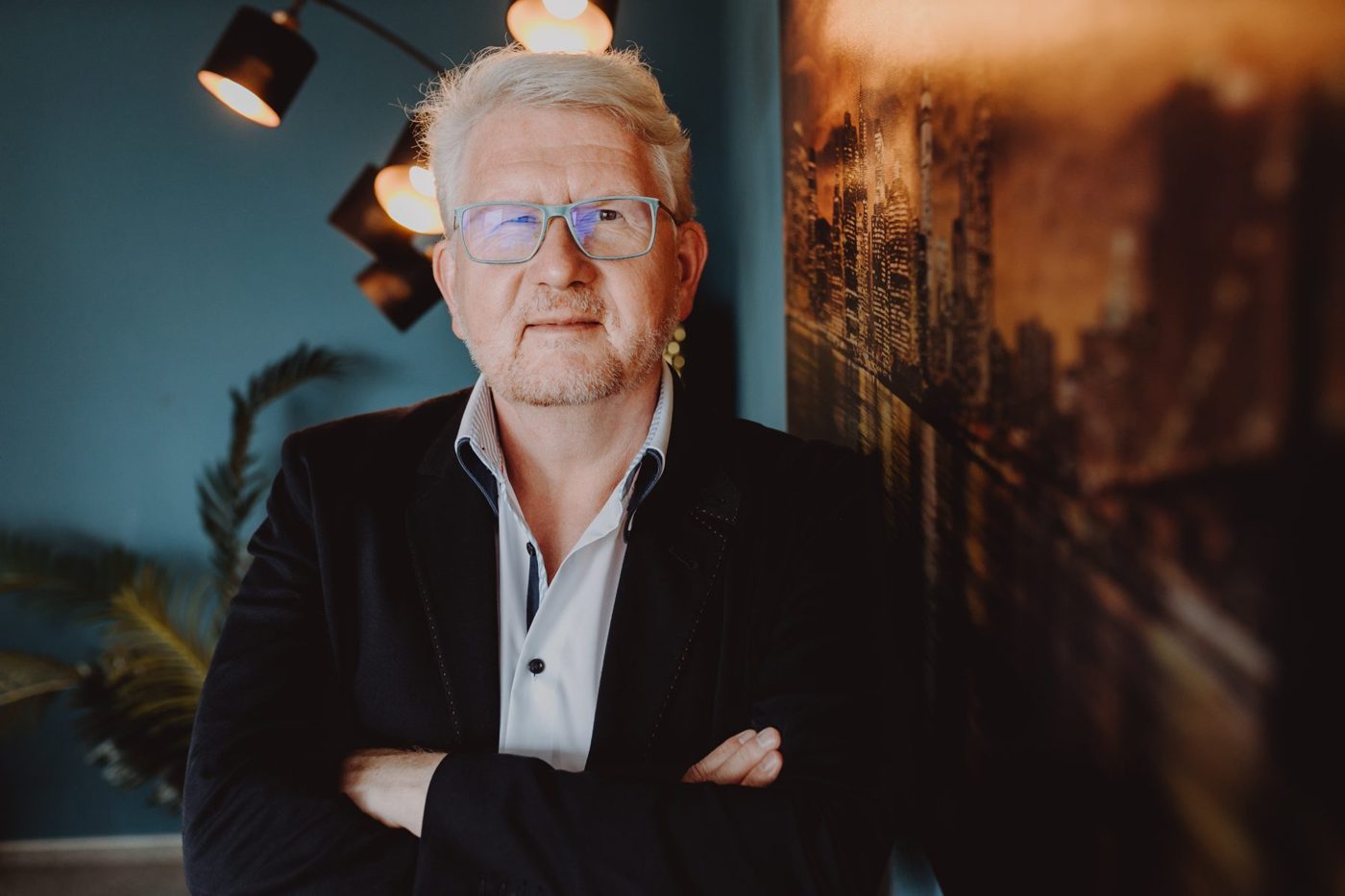 Detlef Gumze mit verschränkten Armen vor petrolfarbener Wand, warmes Licht